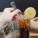 A hand holding a Walco Bosa Nova stainless steel iced tea spoon over a glass of iced tea with a lemon slice.