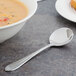 A Walco Meteor stainless steel bouillon spoon next to a bowl of soup on a table.