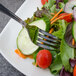 A Walco stainless steel utility fork on a plate of salad with cucumber, tomatoes, and lettuce.