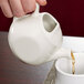 A hand holding a white Tuxton teapot pouring tea into a white cup.