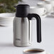 A stainless steel Thermos carafe on a table with cups of coffee.