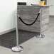 A silver and black rope-style crowd control stanchion with a Lancaster Table & Seating pole.