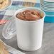 A close-up of a Choice white paper food cup with chocolate ice cream topped with chocolate frosting and a wooden spoon.