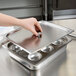 A hand holding a Vollrath metal tray with a lid.
