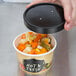 A hand holding a Choice Medley Paper Soup / Hot Food container of fresh hot food with a black lid.