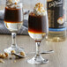 Two glasses of brown liquid with nuts and whipped cream on a table with a bottle of DaVinci Gourmet Sugar Free Hazelnut Flavoring Syrup.