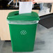 A woman's hand putting paper into a green Lavex recycling bin with a slot lid.
