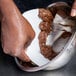 A hand uses an Ateco triangular plastic bowl scraper to mix chocolate frosting in a metal bowl.