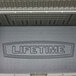 A black metal Lifetime outdoor storage box with rough cut textured metal surfaces and the Lifetime logo on the lid.