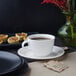 A Villeroy & Boch white porcelain saucer with a cup of coffee on it on a table next to a vase of red flowers.