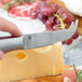 An American Metalcraft stainless steel cheese knife cutting hard cheese on a counter.
