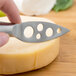 A hand using a Franmara stainless steel cheese knife to cut a wheel of cheese.