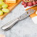 An American Metalcraft stainless steel cheese knife with holes in the blade on a cutting board with grapes.