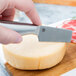 An American Metalcraft stainless steel cheese knife being used to cut a round of hard cheese.