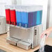 A hand pouring different colored drinks into a Crathco refrigerated beverage dispenser.