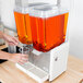 A person pouring orange liquid from a clear plastic cup into a Crathco 5 gallon plastic refrigerated beverage dispenser.