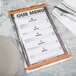 An Alumitique swirl aluminum menu board with orange bands on a table with silverware and a white napkin.