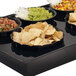 A black fluted bowl filled with tortilla chips on a table with a row of bowls of food.