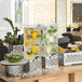A group of Rosseto clear acrylic beverage dispensers with stamped stainless steel bases on a table with fruit and drinks.