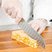 A person in gloves uses a Mercer Culinary Millennia Santoku knife to cut cheese.