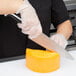 A person in gloves uses a Mercer Culinary Millennia chef knife with a white handle to cut cheese.