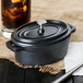 A black Libbey mini cast iron oval Dutch oven with a lid and a glass of liquid.