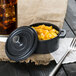 A Libbey mini cast iron pot with a lid on a table with a bowl of macaroni and cheese and a fork.
