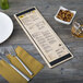 A customizable wood menu board with picture corners on a table with a white plate, silverware, and a glass of liquid.