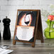A Menu Solutions walnut wood sandwich menu board on a table with a wine glass and a menu.