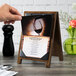 A hand holding a Menu Solutions walnut wood menu board with a picture of a wine glass on a table.