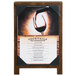 A Menu Solutions walnut wood menu board with picture corners displaying a picture of a wine glass and a bottle of wine on a table.