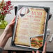 A hand holding a customizable wood menu board with picture corners on a table in a farm-to-table restaurant.