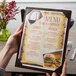A hand holding a Menu Solutions customizable wood menu board with picture corners on a table in a farm-to-table restaurant.