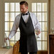 A man in a black apron and tuxedo holding a glass of wine.