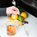 A hand holding a Barfly stainless steel bar spoon and fork next to a glass of amber liquid with ice and a lemon wedge.