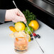 A hand holding a Barfly stainless steel double end stirrer next to a glass of lemonade on a counter.