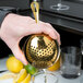 A person holding a Barfly gold-plated Julep strainer over a glass.