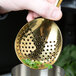 A hand holding a Barfly gold-plated julep strainer over a cocktail with a mint leaf.