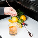 A person using a Barfly copper plated classic bar spoon to stir a glass of amber liquid with ice and a lemon slice.
