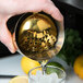 A hand using a Barfly gold-plated scalloped julep strainer to pour liquid into a glass with ice.