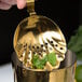 A close-up of a Barfly gold-plated scalloped julep strainer held over a gold cup with a green leaf inside.