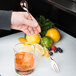 A hand using a Barfly copper plated bar spoon with fork end to stir a cocktail with ice and a lemon wedge.