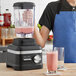 A person in a blue apron using a KitchenAid matte black blender on a wooden surface to make a pink smoothie.