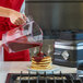 A woman pouring syrup on a stack of pancakes in a plastic container using a KitchenAid blender.