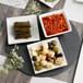 A round black slate tray displaying three square dishes with assorted appetizers.