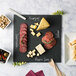 A square black slate tray with labeled sections for cheese and charcuterie, accompanied by a piece of soapstone chalk.