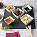 A black square slate tray displaying small white bowls with assorted snacks and dips.