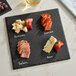 A square black slate tray with four sections labeled for different cheeses and meats, accompanied by soapstone chalk.
