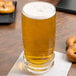 A Libbey Cascade cooler glass of beer on a table with pretzels and a napkin.