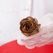 A gloved hand holding a chocolate frosting flower on a stainless steel Ateco flower nail.